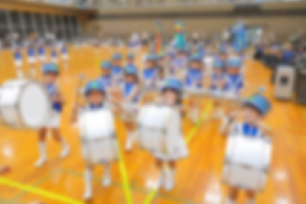 体育館に並ぶ、青い帽子と衣装の鼓笛隊の子どもたち（加工済み）